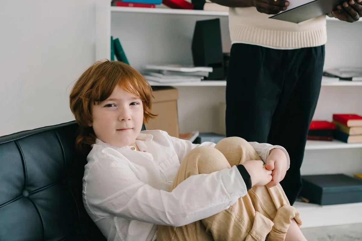 Niño sentado con calma en un sofá durante una sesión de valoración, con un profesional de apoyo visible al fondo sosteniendo una tableta.