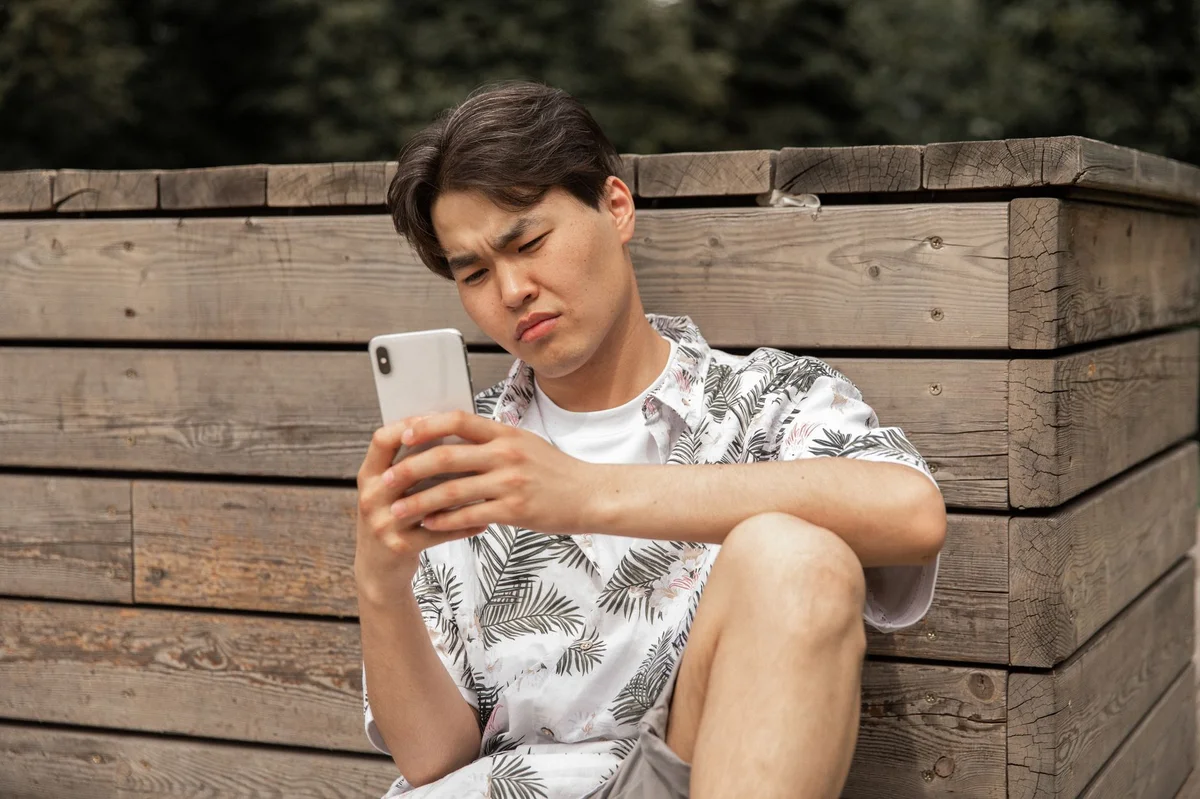 Zaniepokojony mężczyzna z podejrzliwym, zmarszczonym wyrazem twarzy patrzy na ekran smartfona trzymanego w dłoni; ekran telefonu jest zwrócony w jego stronę i nie jest czytelny na zdjęciu.